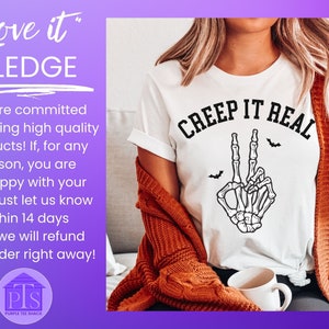 May include: White t-shirt with a black skeleton hand making a peace sign and the text "CREEP IT REAL" in black. The t-shirt is worn by a person with long brown hair and a brown cardigan.