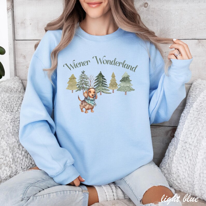 May include: Light blue sweatshirt with the text "Wiener Wonderland" above a watercolor illustration of a dachshund wearing a scarf, surrounded by Christmas trees. The sweatshirt is a solid light blue color.
