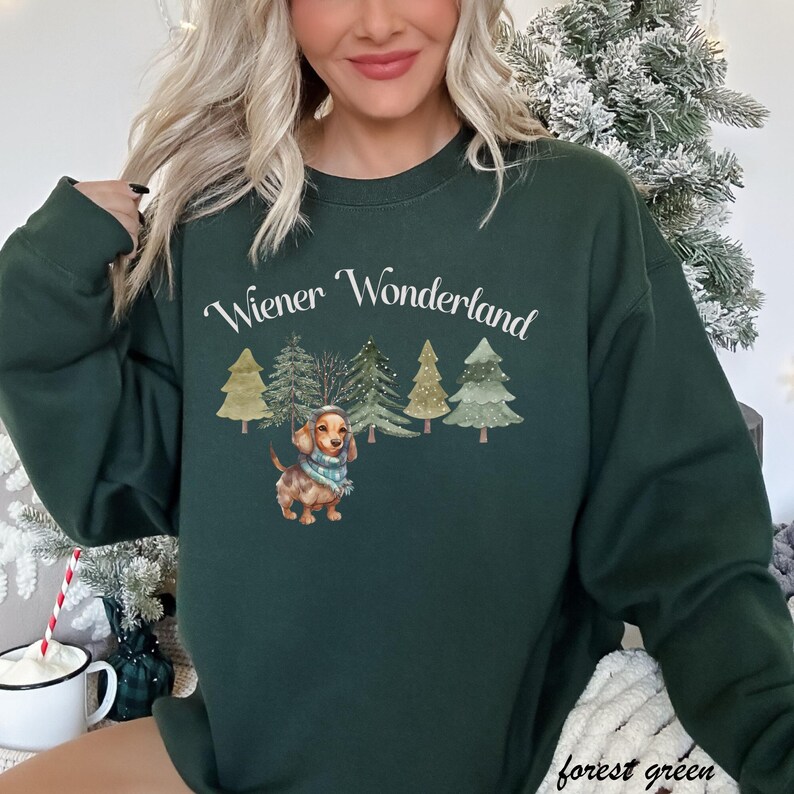 May include: Forest green sweatshirt with the text "Wiener Wonderland" above a watercolor illustration of a dachshund wearing a scarf, surrounded by Christmas trees. The words "forest green" are printed at the bottom.