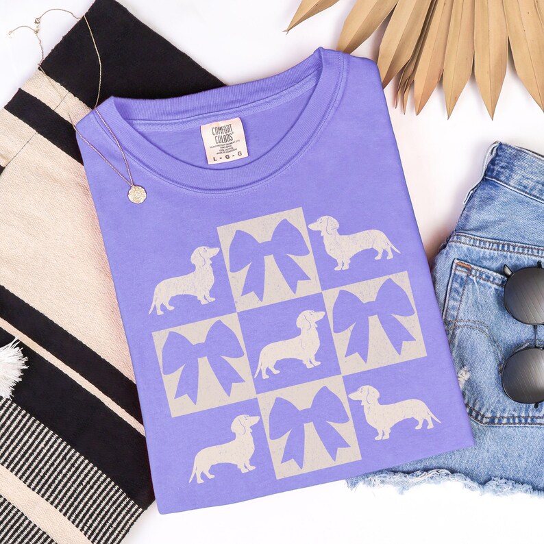 May include: Purple t-shirt with a graphic of a grid of white dachshunds with bows. The bows are beige and the dachshunds are in a grid pattern.