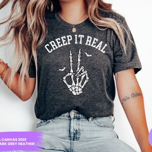 May include: A dark grey heather Bella Canvas 3001 t-shirt with the text "CREEP IT REAL" in white. A white skeleton hand with a peace sign is shown below the text. There are small bats flying around the hand.