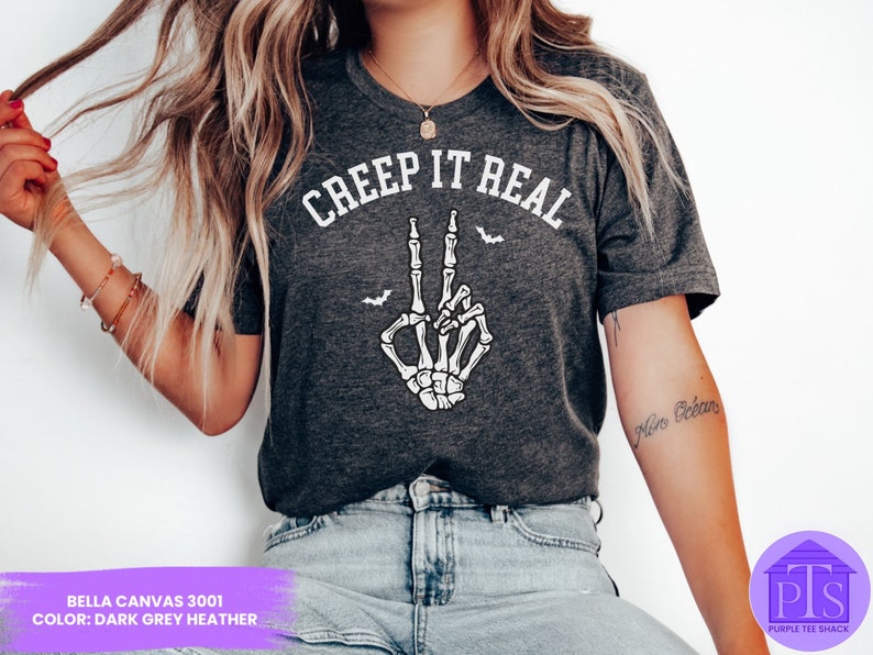 May include: A dark grey heather Bella Canvas 3001 t-shirt with the text "CREEP IT REAL" in white. A white skeleton hand with a peace sign is shown below the text. There are small bats flying around the hand.