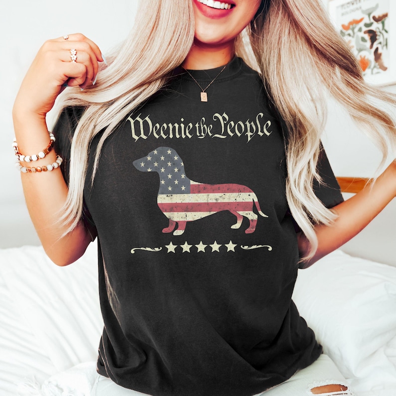May include: Black t-shirt with the text "Weenie the People" above a dachshund silhouette filled with a red, white, and blue American flag design. Below the dog is a row of five stars.