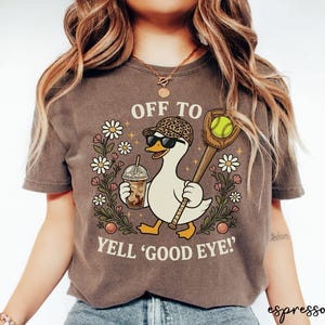 May include: A dark brown t-shirt featuring a graphic of a white duck wearing a leopard print cap and sunglasses, holding an iced coffee and a softball glove with a ball. The text reads OFF TO YELL GOOD EYE surrounded by daisies.