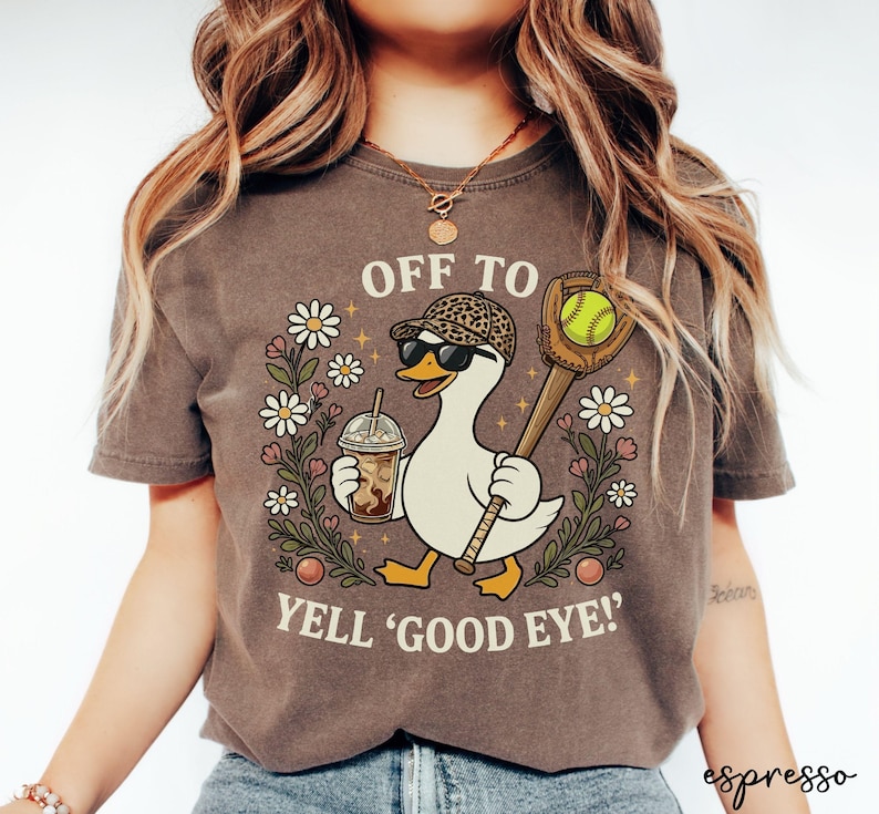 May include: A dark brown t-shirt featuring a graphic of a white duck wearing a leopard print cap and sunglasses, holding an iced coffee and a softball glove with a ball. The text reads OFF TO YELL GOOD EYE surrounded by daisies.