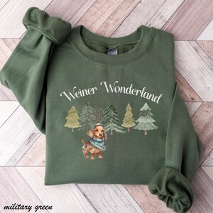 May include: Military green sweatshirt with the text "Weiner Wonderland" above a watercolor illustration of a dachshund wearing a scarf and a hat, with Christmas trees in the background.