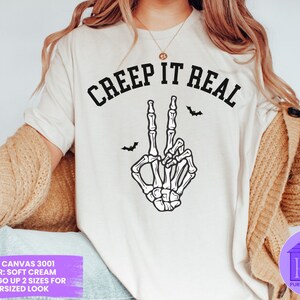 May include: White t-shirt with the text "CREEP IT REAL" in black lettering. A skeleton hand with a peace sign is below the text. The t-shirt is on a model wearing a brown cardigan.