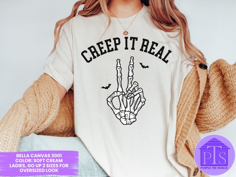 May include: White t-shirt with the text "CREEP IT REAL" in black lettering. A skeleton hand with a peace sign is below the text. The t-shirt is on a model wearing a brown cardigan.