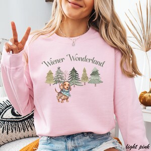 May include: Light pink sweatshirt with the text "Wiener Wonderland" above a watercolor illustration of a dachshund wearing a scarf, surrounded by Christmas trees. The sweatshirt is a solid light pink color.