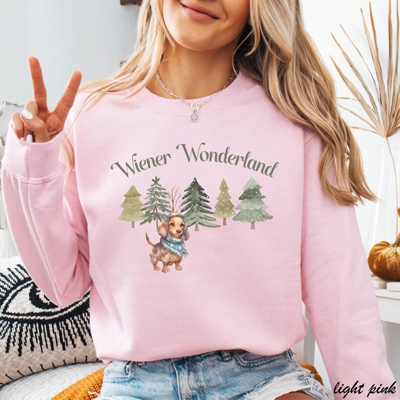 May include: Light pink sweatshirt with the text "Wiener Wonderland" above a watercolor illustration of a dachshund wearing a scarf, surrounded by Christmas trees. The sweatshirt is a solid light pink color.