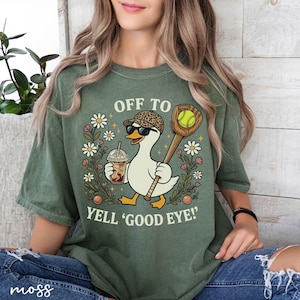 May include: Green t-shirt featuring a cartoon goose in sunglasses and a leopard print hat, carrying a baseball bat and a coffee. The text says "OFF TO YELL 'GOOD EYE!'" with floral accents.