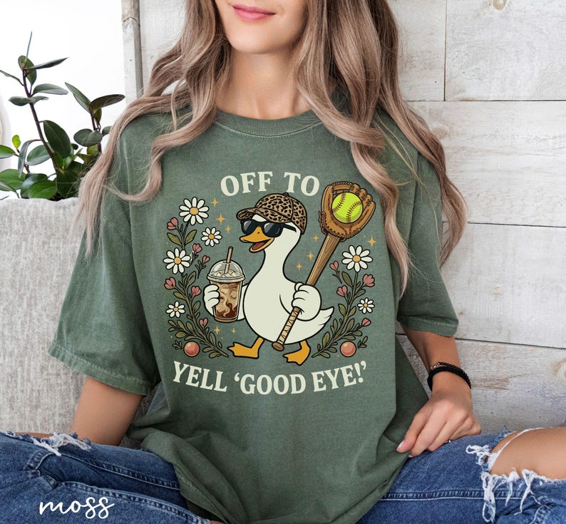 May include: Green t-shirt featuring a cartoon goose in sunglasses and a leopard print hat, carrying a baseball bat and a coffee. The text says "OFF TO YELL 'GOOD EYE!'" with floral accents.