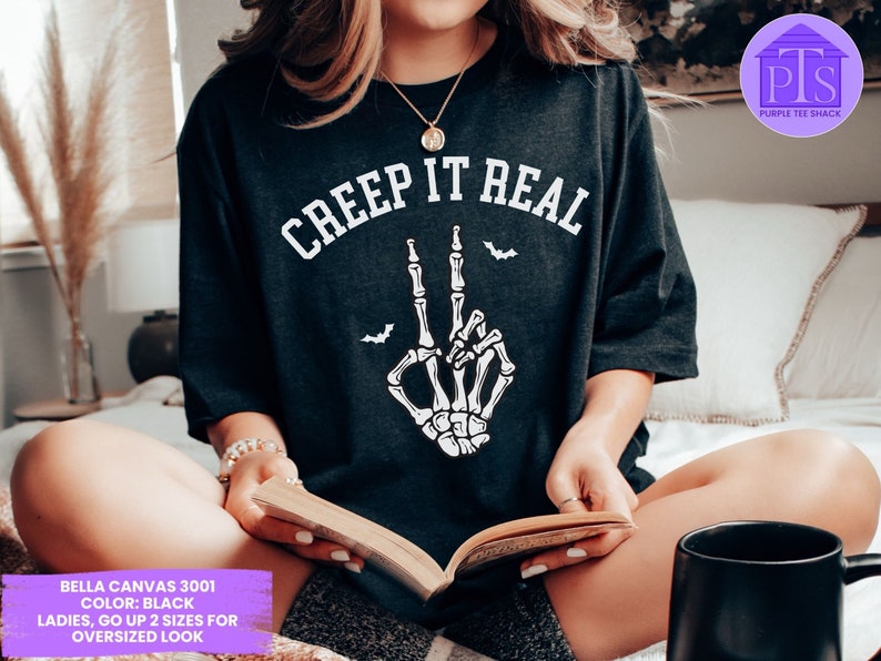 May include: Black t-shirt with the text "CREEP IT REAL" in white lettering. A white skeleton hand with two fingers up is in the center of the shirt. The shirt is being worn by a person sitting on a bed.