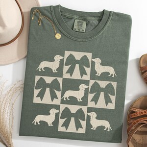 May include: A green t-shirt with a white graphic of a checkered pattern featuring dachshunds and bows. The text "COMFORT COLORS" is on the tag.