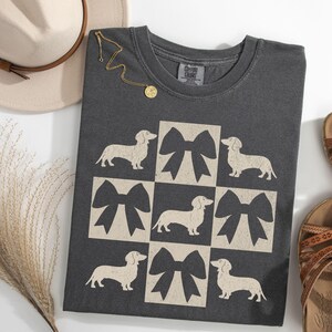 May include: A dark gray t-shirt with a graphic design of white dachshunds and black bows in a checkered pattern. The shirt has a crew neck and short sleeves.
