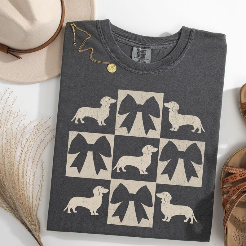 May include: A dark gray t-shirt with a graphic design of white dachshunds and black bows in a checkered pattern. The shirt has a crew neck and short sleeves.