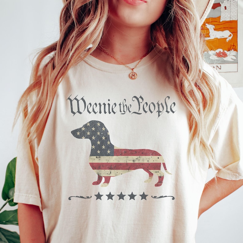 May include: Cream-colored t-shirt featuring a dachshund silhouette filled with an American flag design. The text "Weenie the People" is arched above the dog, with five stars below. A patriotic and playful design.