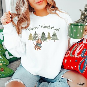 May include: White long-sleeve sweatshirt with the text "Wiener Wonderland" above a watercolor illustration of a dachshund wearing a scarf, with Christmas trees in the background. A green mug with white tree designs is held in the hand.