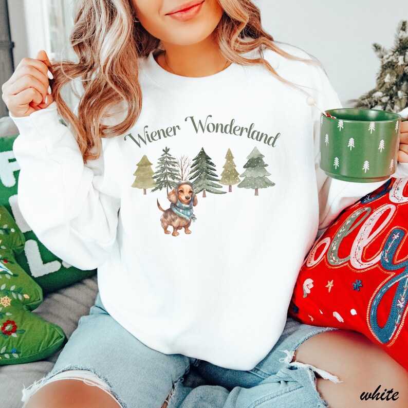May include: White long-sleeve sweatshirt with the text "Wiener Wonderland" above a watercolor illustration of a dachshund wearing a scarf, with Christmas trees in the background. A green mug with white tree designs is held in the hand.