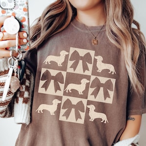 May include: A brown t-shirt with a graphic of a repeated pattern of dachshunds and bows. The graphic is in a cream color.