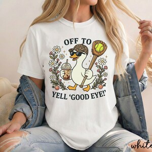 May include: A white t-shirt featuring a graphic of a duck wearing a leopard print baseball cap and sunglasses, holding an iced coffee and a baseball bat with a glove. The text reads OFF TO YELL 'GOOD EYE!' surrounded by flowers.