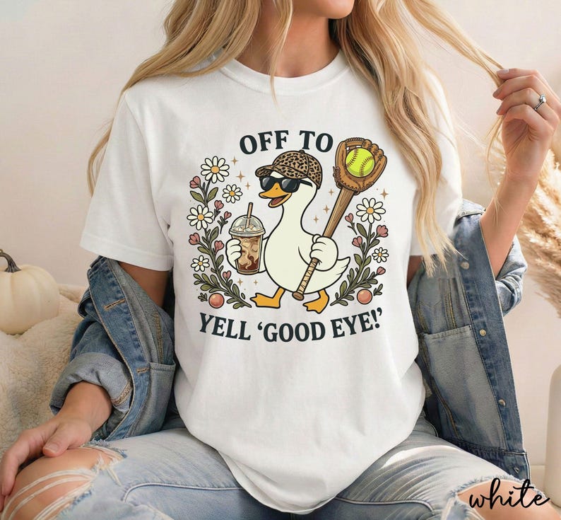 May include: A white t-shirt featuring a graphic of a duck wearing a leopard print baseball cap and sunglasses, holding an iced coffee and a baseball bat with a glove. The text reads OFF TO YELL 'GOOD EYE!' surrounded by flowers.