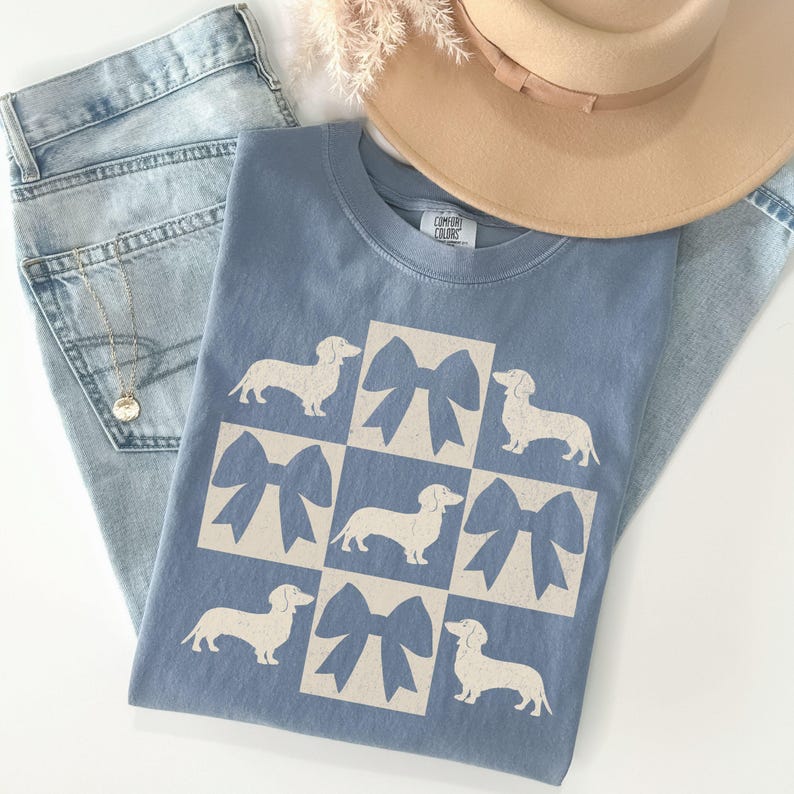 May include: A light blue t-shirt with a white graphic of a checkered pattern featuring dachshunds and bows. The shirt has the text "COMFORT COLORS" on the neck.