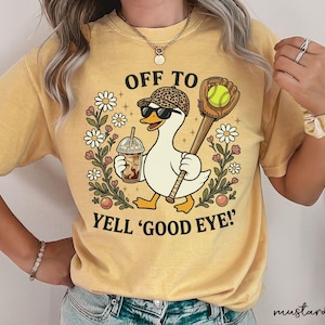 May include: A mustard yellow t-shirt featuring a graphic of a white duck wearing a leopard print baseball cap and sunglasses. The duck holds an iced coffee and a baseball glove with a ball. Text reads OFF TO YELL 'GOOD EYE!' surrounded by daisies.