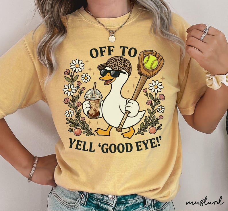 May include: A mustard yellow t-shirt featuring a graphic of a white duck wearing a leopard print baseball cap and sunglasses. The duck holds an iced coffee and a baseball glove with a ball. Text reads OFF TO YELL 'GOOD EYE!' surrounded by daisies.