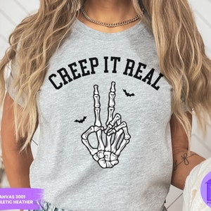 May include: A gray t-shirt with the text "CREEP IT REAL" in black lettering. A skeleton hand with a peace sign gesture is below the text. Two black bats are above the text.