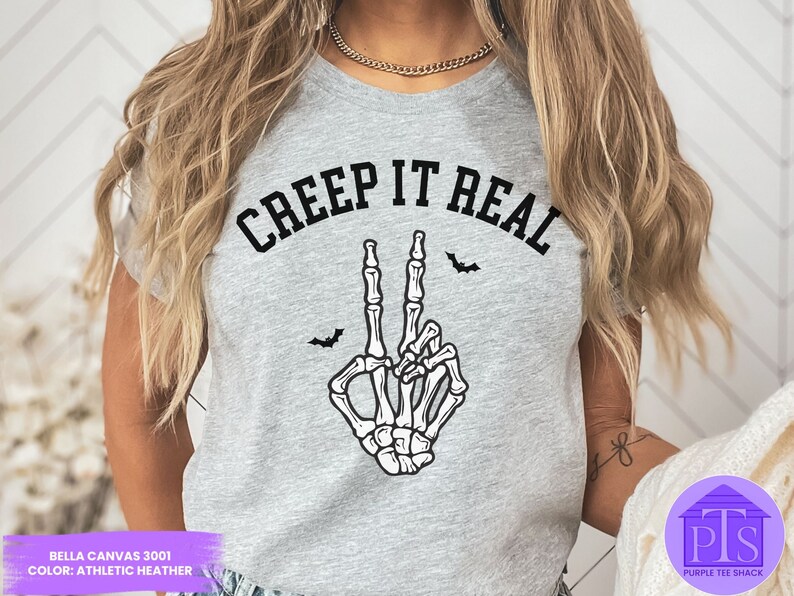 May include: A gray t-shirt with the text "CREEP IT REAL" in black lettering. A skeleton hand with a peace sign gesture is below the text. Two black bats are above the text.