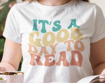 May include: White t-shirt with the text "IT'S A GOOD DAY TO READ" in a retro font with a rainbow colour scheme. The text is in a large font and is centred on the shirt.