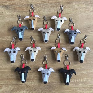 May include: A collection of twelve felt dog head keychains, each featuring a distinct coat colour including black, grey, tan, and cream. Every keychain has a white felt blaze on the snout, dark button eyes, and a red ribbon attached to a metal ring.