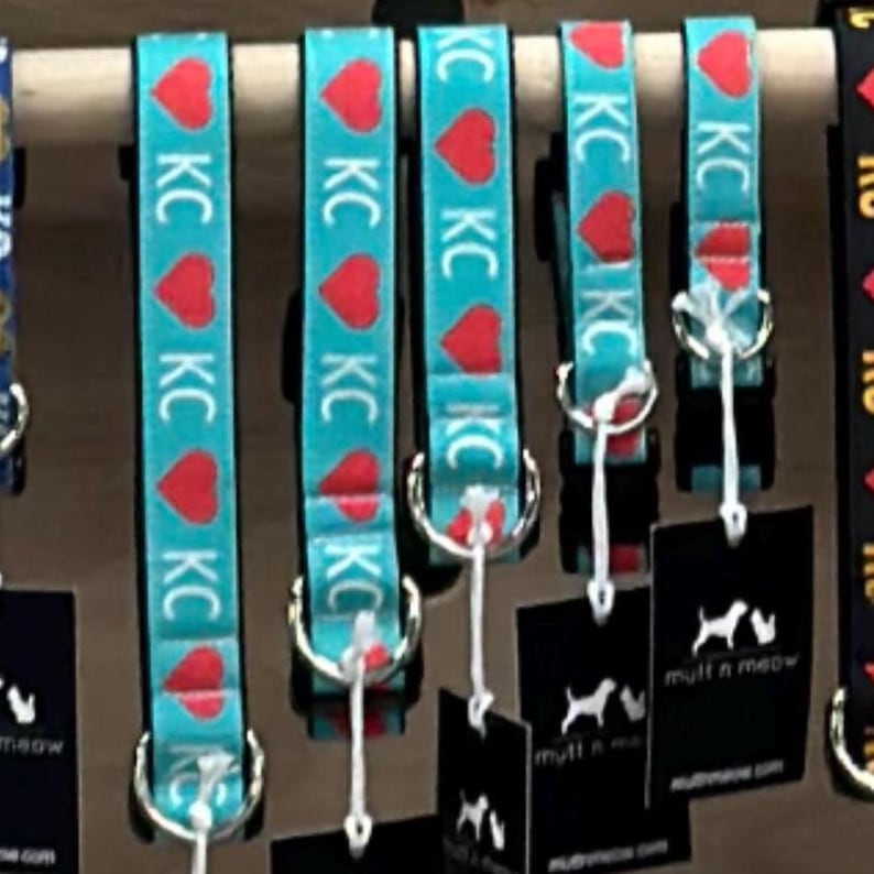 May include: Turquoise dog collars with red heart and "KC" designs are displayed. Each collar has a metal ring and a white tag. The collars are hanging on a wooden dowel. Black tags with a dog silhouette and the words "mutt n meow" are attached.