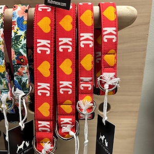 May include: Red dog collars with yellow heart patterns and the letters "KC" in white. The collars are displayed on a wooden rack, with tags hanging below. Other floral patterned collars are also visible.