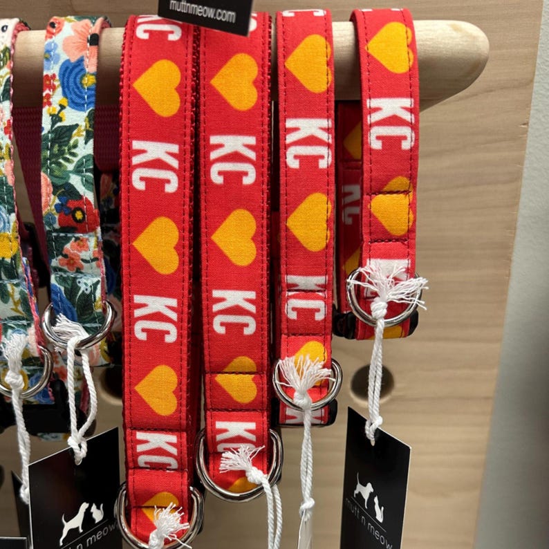 May include: Red dog collars with yellow heart patterns and the letters "KC" in white. The collars are displayed on a wooden rack, with tags hanging below. Other floral patterned collars are also visible.