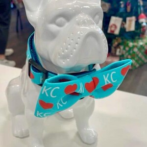 May include: A white ceramic French bulldog statue wearing a turquoise dog collar and bow tie. The bow tie has red hearts and the letters "KC" printed on it. The dog collar is turquoise with a black buckle.