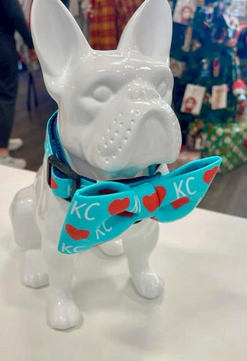 May include: A white ceramic French bulldog statue wearing a turquoise dog collar and bow tie. The bow tie has red hearts and the letters "KC" printed on it. The dog collar is turquoise with a black buckle.