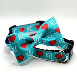 May include: A turquoise dog collar and bow tie set. The collar and bow tie feature a repeating pattern of red hearts and the letters "KC" in white. The collar has a black buckle and a silver metal loop.