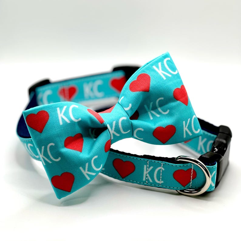May include: A turquoise dog collar and bow tie set. The collar and bow tie feature a repeating pattern of red hearts and the letters "KC" in white. The collar has a black buckle and a silver metal loop.