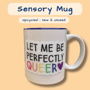 May include: White ceramic mug with a blue rim. The mug has a rainbow sticker that says "Let me be perfectly queer" with a purple heart.