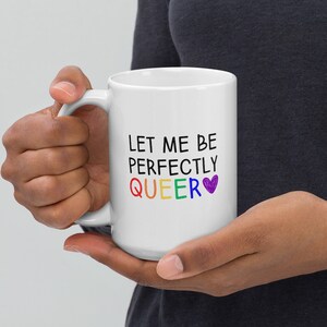 May include: White ceramic coffee mug with the text "Let me be perfectly QUEER" in black lettering with a rainbow colored "QUEER" and a purple heart.