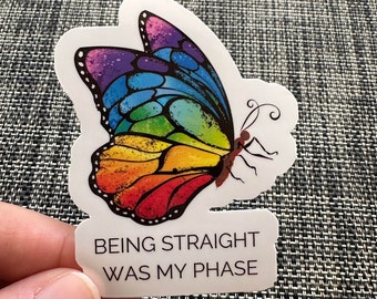Può includere: Un adesivo con una farfalla colorata con ali arcobaleno. La farfalla ha un corpo marrone e contorni neri. Il testo "BEING STRAIGHT WAS MY PHASE" è sotto la farfalla.