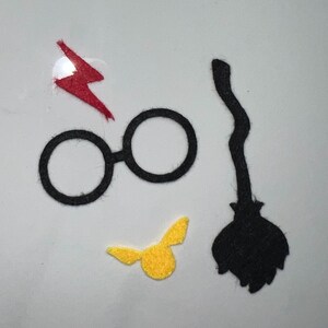 May include: A set of four felt Harry Potter themed cutouts. The set includes a pair of round glasses, a lightning bolt, a golden snitch, and a black broom.