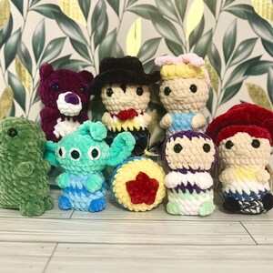Puede incluir: Una colecci&oacute;n de juguetes de peluche tejidos a crochet, que incluye un dinosaurio verde, un extraterrestre azul, un oso rojo, un personaje morado y verde, un personaje rojo y amarillo, un vaquero, una princesa y un personaje rojo y amarillo con un sombrero rojo.
