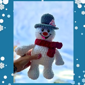 May include: A crocheted snowman toy with a red scarf, a grey hat with a pink flower, and a white body. The snowman has a red nose, black eyes, and a smiling mouth with a pink tongue sticking out. The snowman is being held in a hand against a blue sky background with white snowflakes.