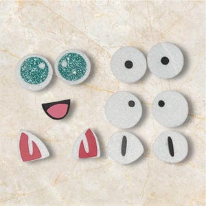 Könnte beinhalten: Eine Sammlung von Bastelmaterialien, darunter zwei glitzernde, türkisfarbene Augen, mehrere weiße Augen mit schwarzen Pupillen, ein rosa Mund und zwei rot umrandete Ohren. Für Bastel- und DIY-Projekte.