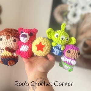 Puede incluir: Una colecci&oacute;n de personajes de juguetes de crochet de la pel&iacute;cula Toy Story. Los personajes incluyen a Woody, Jessie, Buzz Lightyear, Rex y un extraterrestre verde. Los juguetes est&aacute;n hechos de hilo de colores y tienen ojos saltones. Los juguetes est&aacute;n dispuestos en fila sobre una mano.