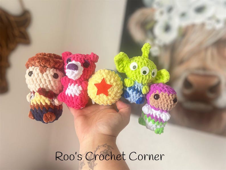Puede incluir: Una colecci&oacute;n de personajes de juguetes de crochet de la pel&iacute;cula Toy Story. Los personajes incluyen a Woody, Jessie, Buzz Lightyear, Rex y un extraterrestre verde. Los juguetes est&aacute;n hechos de hilo de colores y tienen ojos saltones. Los juguetes est&aacute;n dispuestos en fila sobre una mano.