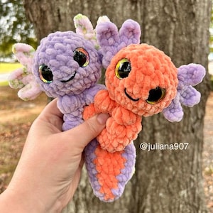 Peut inclure: Deux peluches axolotl crochetées à la main. L'une est violette avec des accents roses et verts, l'autre est orange avec des accents violets. Les deux ont de grands yeux irisés et des visages souriants. Les peluches sont douces et câlines, parfaites pour un jouet d'enfant ou une décoration de chambre.